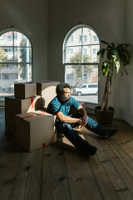 A man with curly hair, wearing a blue T-shirt and dark pants, is seated on a wooden floor inside a room with two large arched windows. Natural daylight illuminates the space, which contains several cardboard boxes sealed with red tape, stacked around him, some on the floor and partially resting against furniture. A tall potted plant with broad leaves is positioned near the right window. The scene depicts a home relocation process, with the man appearing to rest during packing or moving activities. The room has a minimalistic interior, and the designated service is related to removals, as indicated by the website domain manwithvancatford.co.uk and the page topic about tight access moves in Catford.
