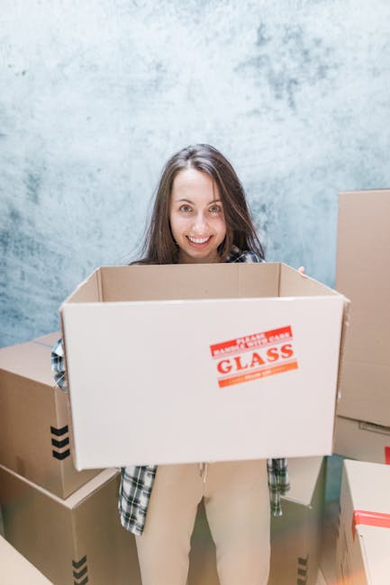 Close-up of three cardboard moving boxes placed on a wooden floor inside a property. The top box, positioned slightly behind the others, has the word 'STUFF' handwritten in red marker on its side, along with a small smiley face. The middle box prominently displays the word 'CLOTHES' handwritten in black marker. The boxes appear to be packed with household items, ready for home relocation. Surrounding the boxes are additional packing parcels, and the environment suggests an interior space in the process of being prepared for moving or packing. This scene reflects the packing stage of a furniture transport or removal service by Man with Van Catford, highlighting careful organisation of belongings before loading the vehicle for transportation to a new location.