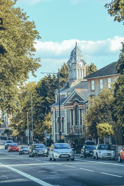 A view of a busy street in a residential area in Catford during daytime, featuring several parked and moving cars along the road. In the background, a historic building with a clock tower and ornate a